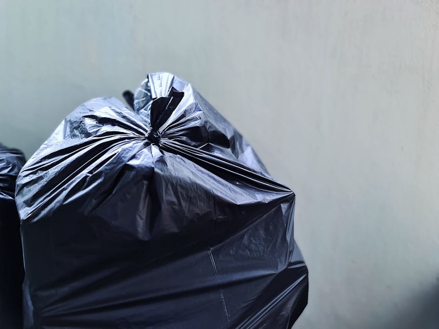 Close-up view of several black plastic rubbish bin liners filled with waste, with the top of the liners tied securely. The liners are made of glossy, slightly crinkled plastic material, reflecting lig